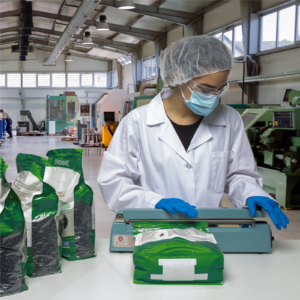 Mujer en un entorno de fábrica, usando equipo de protección personal y sellando varias bolsas grandes sellables de frijoles negros orgánicos con la selladora manual por impulso.