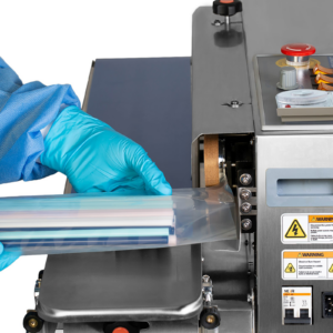 Una persona que lleva guantes desechables y ropa utiliza la selladora de banda JORES TECHNOLOGIES® para sellar una bolsa de plástico transparente llena de pajitas. La máquina está colocada en posición horizontal mientras la bolsa se desplaza apoyada sobre la banda giratoria.