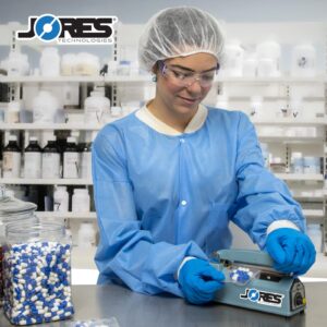 Mujer en un entorno de laboratorio con una variedad de frascos de medicamentos en estantes. Lleva una red desechable para el cabello, bata, gafas y guantes, y está operando la selladora manual por impulso JORES TECHNOLOGIES® de 4 pulgadas para sellar una bolsa con varias píldoras azules y blancas