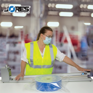 Mujer en un entorno de fábrica usando EPP y sellando una bolsa que contiene un gran rollo de cable azul con una selladora manual de impulso para bolsas extra larga JORES TECHNOLOGIES®.
