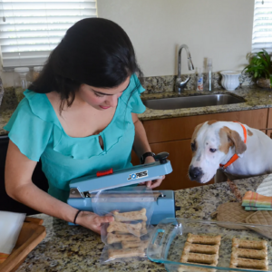 Mujer y su perro en su cocina. Ella está empaquetando premios para perros en bolsas sellables con la selladora manual por impulso JORES TECHNOLOGIES® de 8 pulgadas.