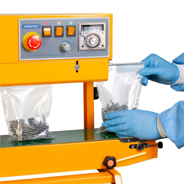 Trabajador con equipo de protección personal introduciendo bolsas de plástico llenas de clavos en la selladora continua de bolsas con calor JORES TECHNOLOGIES® amarilla. La selladora de bolsas está colocada para aplicaciones verticales