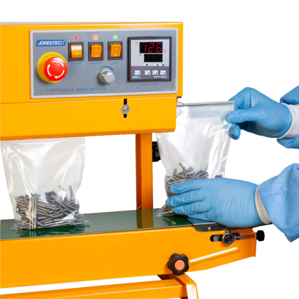 Un operador con guantes azules y ropa desechable insertando bolsas de plástico llenas de clavos en la selladora continua de banda amarilla JORES TECHNOLOGIES®. La selladora de bolsas está posicionada para aplicaciones verticales