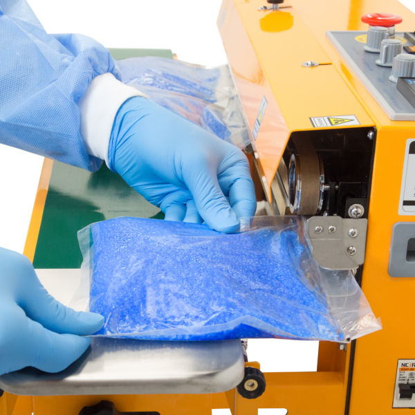operador con guantes azules insertando una bolsa de plástico llena de perlas de cera azules en una selladora continua de banda horizontal y vertical JORES TECHNOLOGIES®. La selladora de bolsas está posicionada para aplicaciones horizontales