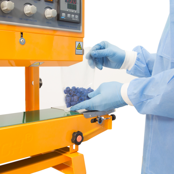 Un operador que lleva guantes azules y ropa desechable insertando una bolsa de plástico llena de piezas plásticas azules en la selladora continua de banda amarilla JORES TECHNOLOGIES®. La selladora de bolsas está posicionada para aplicaciones verticales