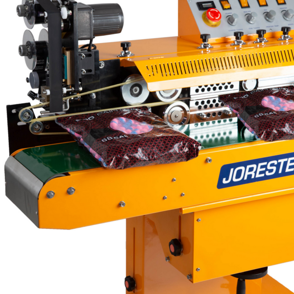 primer plano de la selladora de banda continua horizontal autoportante JORES TECHNOLOGIES® sellando y codificando por estampado en caliente bolsas de plástico llenas de bolas azules y rosas. También muestra el codificador de estampado en caliente de la selladora de banda continua con un rollo de cinta de tinta negra para estampado en caliente instalado
