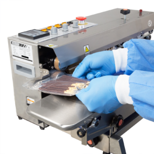 Las manos de una persona que lleva ropa desechable de EPP mientras sella bolsas de plástico llenas de galletas con chocolate con una selladora continua de banda JORES TECHNOLOGIES® de acero inoxidable, de izquierda a derecha, configurada para aplicaciones horizontales.