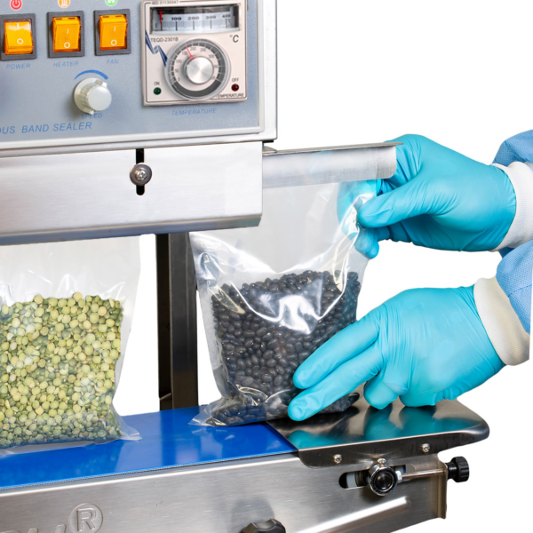 Un operador con guantes azules insertando bolsas transparentes con frijoles negros y lentejas verdes en una selladora continua de banda JORES TECHNOLOGIES® para cerrar las bolsas