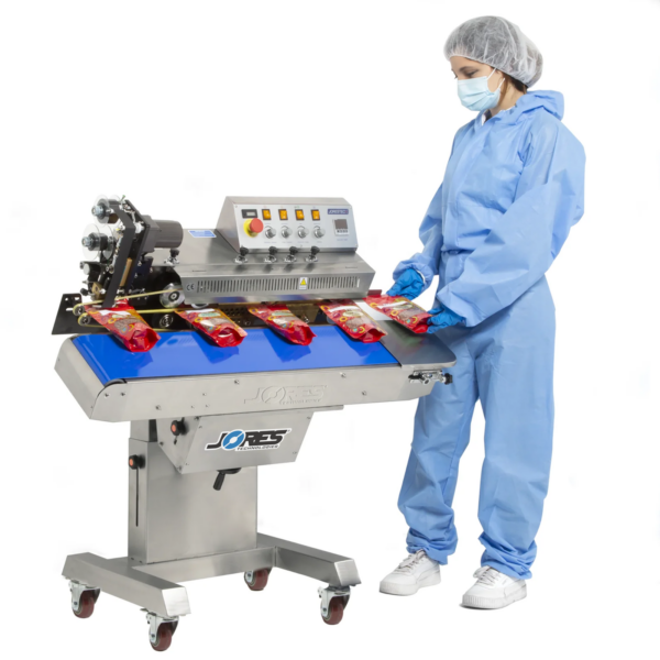 Operador con overol azul insertando una bolsa de plástico roja para ser sellada y codificada por una selladora continua de banda JORES TECHNOLOGIES® de acero inoxidable. Toda la máquina, excepto la base, está inclinada hacia adelante, mostrando que la máquina puede ajustarse a una posición horizontal o inclinada.
