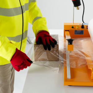 Un trabajador utilizando la unidad de embalaje retráctil para sellar unas cajas. Una pistola de calor está montada en el soporte de pistola de la máquina y un rollo de film descansa sobre las barras portarrollos.