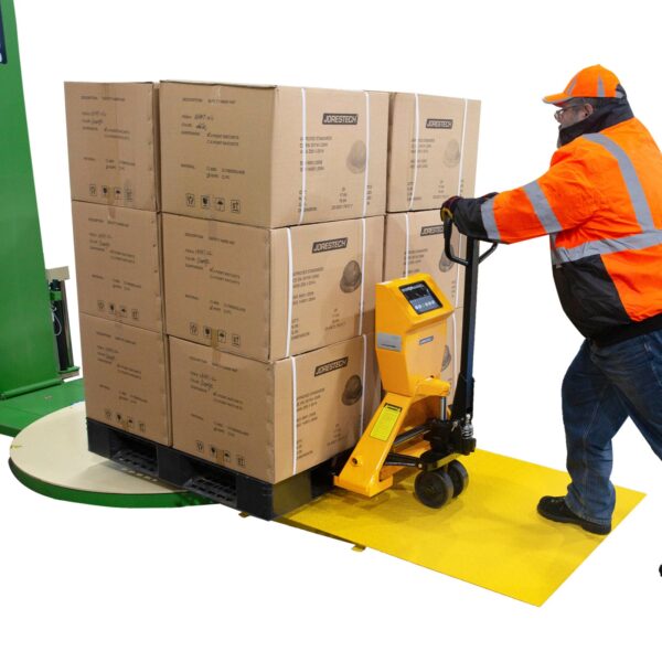 Hombre con una chaqueta de seguridad naranja de alta visibilidad colocando un palé cargado de cajas sobre la plataforma giratoria del transpalet. Está pisando la rampa de carga y parte del transpalet también está bajando por la rampa de carga.