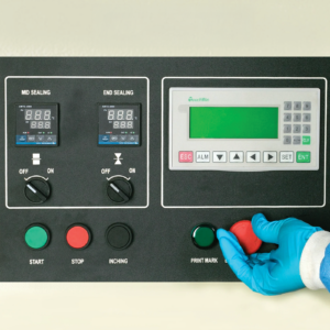 Se ve a un operador que lleva un guante desechable de EPP azul utilizando una máquina envasadora horizontal HFFS flow pack. Se muestra el panel de control y está a punto de presionar el botón de parada de emergencia.