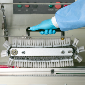 Primer plano de las piezas internas de una máquina de envasado Flow JORES TECHNOLOGIES®® y la mano de una persona abriendo una puerta transparente para inspeccionar los cepillos barrenderos