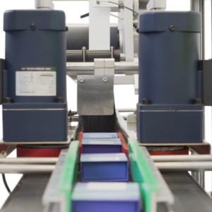 El etiquetador automático en producción aplicando etiquetas sobre la parte superior de pequeñas cajas planas azules colocadas en la cinta transportadora del aplicador automático de etiquetas JORES TECHNOLOGIES®