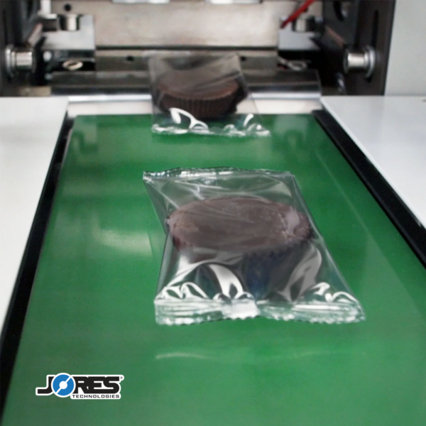 Paquete tipo almohada con un producto comestible en el interior, sobre una cinta transportadora verde de salida para llevar después de ser sellado por una máquina automática de envasado flow pack HFFS