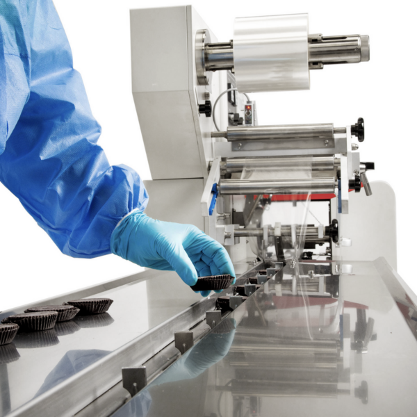 Primer plano de un operador con guantes azules colocando un producto comestible sobre la cinta transportadora de cadena plana de una máquina flow pack de JORES TECHNOLOGIES®. Se pueden ver las barras empujadoras mientras coloca un producto justo delante de cada barra empujadora.