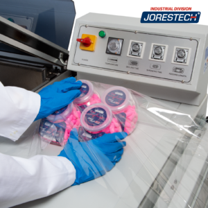 Primer plano de la cámara de retractilado o de un sistema de envoltura retráctil JORES TECHNOLOGIES® y una persona colocando 4 envases de tamaño mediano dentro de un material de poliolefina centerfold antes de introducirlos en la cámara de termorretracción para formar un paquete.