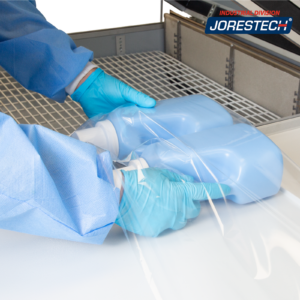 Primer plano de la cámara de retracción de un sistema de envoltura retráctil JORES TECHNOLOGIES® que muestra las manos de una persona insertando 2 envases en la película plegada al centro antes de colocar el paquete a retraer dentro de la cámara de termorretracción.