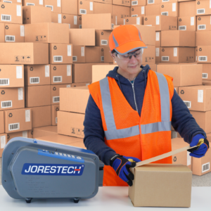 Un trabajador cerrando cajas usando el dispensador de cinta de papel engomada JORES TECHNOLOGIES®. El dispensador de cinta de mesa está sobre una mesa de trabajo junto con la caja que se está cerrando. El fondo está lleno de cajas de cartón marrones cerradas con cinta engomada