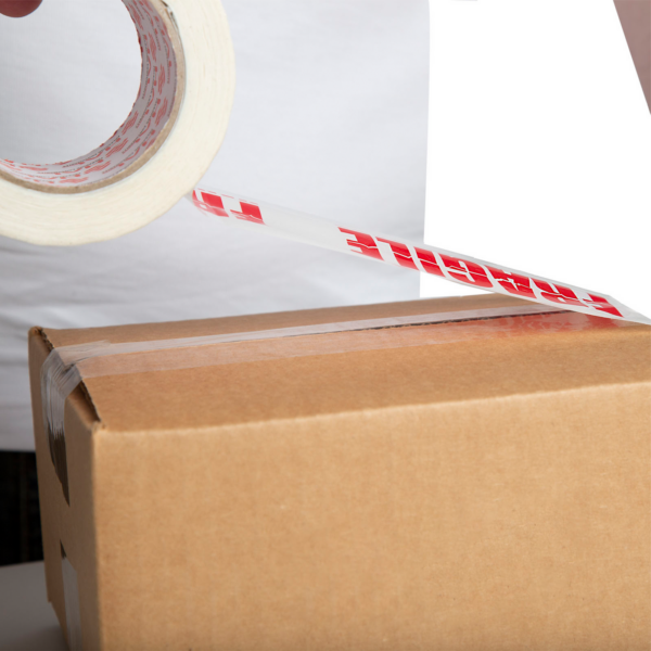 persona con camisa blanca usando cinta blanca impresa "FRAGILE" para cerrar una caja marrón