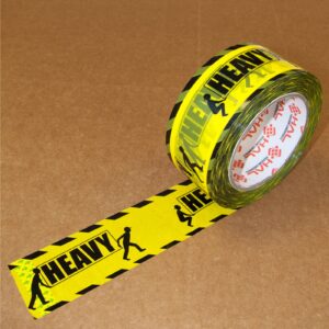 cinta de embalaje impresa en amarillo y negro con dos figuras de hombres sosteniendo un cartel que dice heavy sobre la cinta encima de una caja marrón