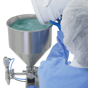 Persona con ropa y guantes desechables de pie junto a una llenadora de pistón. Está dispensando una crema verde en la tolva de una llenadora de pistón de sobremesa JORES TECHNOLOGIES® para pasta