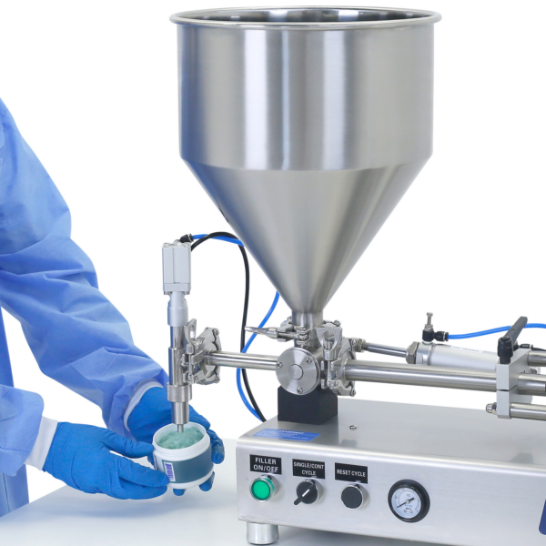 Una persona con ropa de EPP y guantes desechables de pie frente a la boquilla de una llenadora de pistón. Está retirando un recipiente lleno de un gel viscoso azul después de haber sido llenado por la llenadora de pistón de acero inoxidable JORES TECHNOLOGIES® colocada sobre una mesa de trabajo.