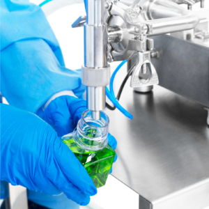 Una persona que lleva guantes de seguridad de nitrilo operando el Llenador de Pistón para Baja Viscosidad de JORES TECHNOLOGIES®. El operario está posicionando una botella de 100 ml debajo de la boquilla antigoteo para llenarla con precisión mediante la máquina de llenado de líquidos.