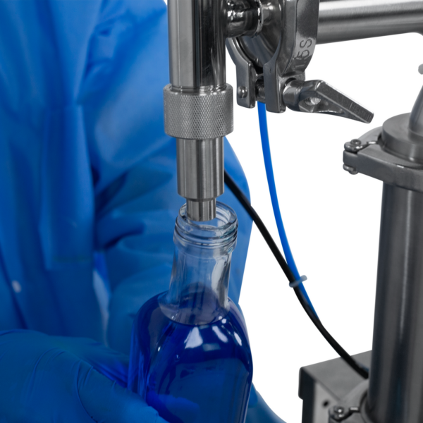 Trabajador con guantes de seguridad de nitrilo operando la llenadora de pistón para líquidos de baja viscosidad JORES TECHNOLOGIES® para botellas de 1000 ml. La persona está colocando una botella de 1000 ml debajo de la punta de la boquilla antigoteo para llenarla con precisión mediante la máquina llenadora de líquidos.