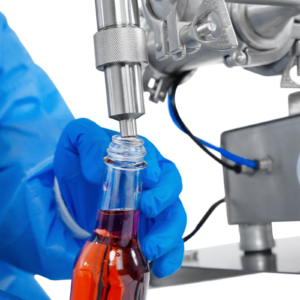 Trabajador con guantes de seguridad de nitrilo operando el pistón dosificador de baja viscosidad JORES TECHNOLOGIES®. La persona está colocando una botella de 250 ml debajo de la punta de la boquilla antigoteo para llenarla con precisión mediante la máquina llenadora de líquidos.