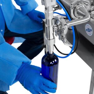 Un operario con guantes de seguridad de nitrilo operando el llenador de pistón para baja viscosidad JORES TECHNOLOGIES®. La persona está posicionando una botella de 500 ml debajo de la punta de la boquilla antigoteo para ser llenada con precisión por la máquina llenadora de líquidos.