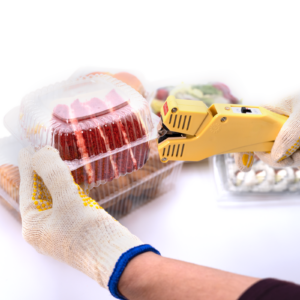 Trabajador usando una selladora manual de calor directo para clamshell para cerrar varias cajas transparentes llenas de verduras, galletas, sushi, pastel, etc.