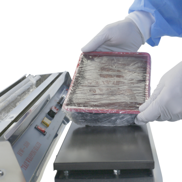 operador con guantes blancos envolviendo una bandeja de alimentos con film retráctil usando una envolvedora manual de bandejas