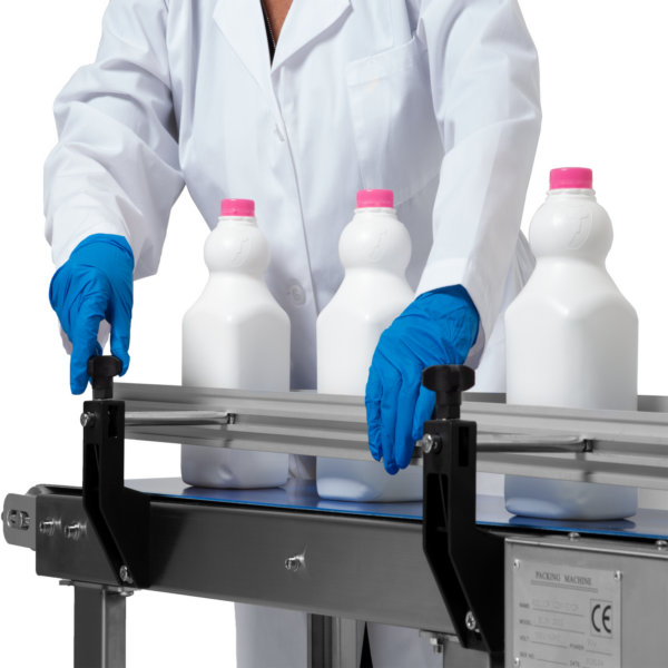 operario con bata de laboratorio blanca y guantes azules ajustando el riel lateral en una cinta transportadora motorizada JORES TECHNOLOGIES® con botellas de leche blancas