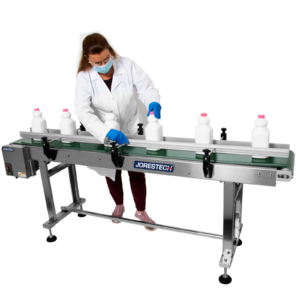 mujer morena con bata de laboratorio blanca y guantes azules ajustando botellas de leche blancas en una cinta transportadora motorizada con estructura de acero y banda verde que incluye barandillas laterales de protección