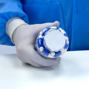 Persona con guantes de nitrilo sosteniendo un envase cerrado con un liner de sellado por inducción con pestañas. El envase está lleno de tabletas medicinales azules y blancas.