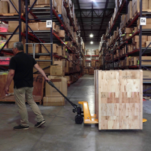 Un trabajador tirando de un transpaleta amarillo y negro JORES TECHNOLOGIES® en un almacén lleno de cajas. El transpaleta tiene varias cajas grandes y pesadas de madera encima