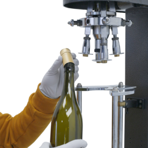 operario con camisa amarilla y guantes blancos insertando una tapa metálica en la botella antes de cerrarla con la encapsuladora de mesa gris JORES TECHNOLOGIES®