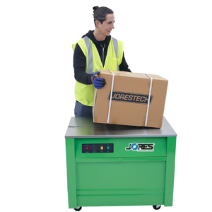 Trabajador operando una flejadora semiautomática de polipropileno verde JORES TECHNOLOGIES® con ruedas. Tiene una caja de cartón colocada sobre la máquina con 3 flejes de polipropileno asegurando la caja alrededor