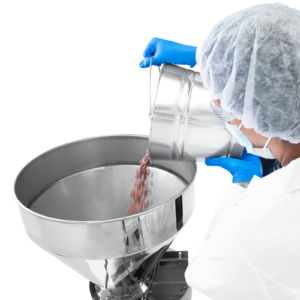 Una persona que lleva guantes de nitrilo azules y un conjunto de equipos de protección personal vierte un cubo lleno de frijoles en la tolva de acero inoxidable de la llenadora volumétrica semiautomática JORES TECHNOLOGIES®