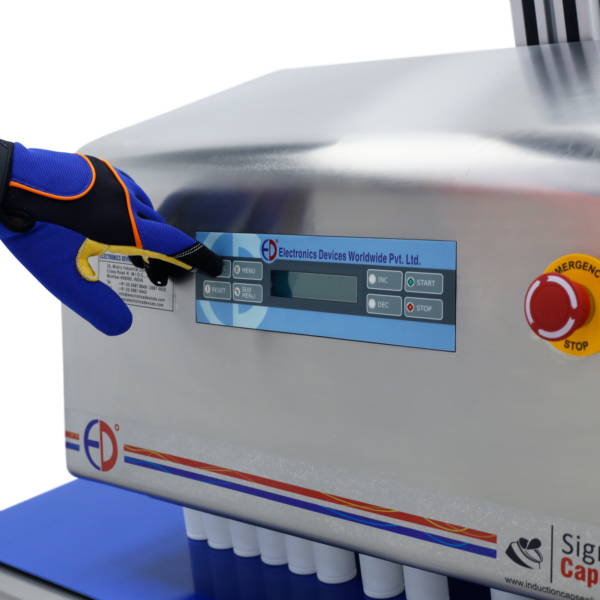 Se puede ver a un operador con un guante azul presionando un botón en el panel de control de una máquina selladora por inducción SIGMA.