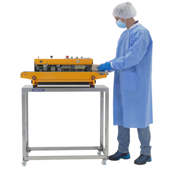 Una persona que lleva EPP desechable y guantes de nitrilo utilizando una selladora continua de banda, colocada sobre una mesa de trabajo de preparación de acero inoxidable JORES TECHNOLOGIES®