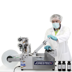 Un hombre que lleva equipo de protección personal. Está operando la etiquetadora semiautomática JORES TECHNOLOGIES® aplicando etiquetas a recipientes redondos. Se muestra un conjunto de 3 botellas con etiquetas impresas que contienen información escrita