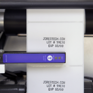 primer plano del sensor y la etiqueta impresa con el nombre de la marca, número de lote y fecha de producción en un aplicador de etiquetas semiautomático con impresora integrada