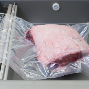 Bolsa sellada al vacío con un gran trozo de carne en su interior. La bolsa está colocada dentro de la cámara de la selladora al vacío comercial de sobremesa JORES TECHNOLOGIES® de una sola cámara, de acero inoxidable, con doble barra de sellado