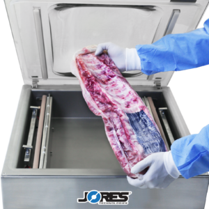 Persona con guantes y ropa desechables. Sostiene una gran parrilla de costillas después de haber sido envasada al vacío por la selladora al vacío comercial de sobremesa JORES TECHNOLOGIES® de cámara única de acero inoxidable con doble barra de sellado en el fondo.