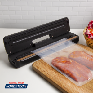 Máquina de sellado al vacío negra JORES TECHNOLOGIES® para uso doméstico sobre una encimera de cocina. Una bolsa de vacío gofrada llena de salami reposa sobre una tabla de madera junto a la máquina selladora, lista para ser envasada.