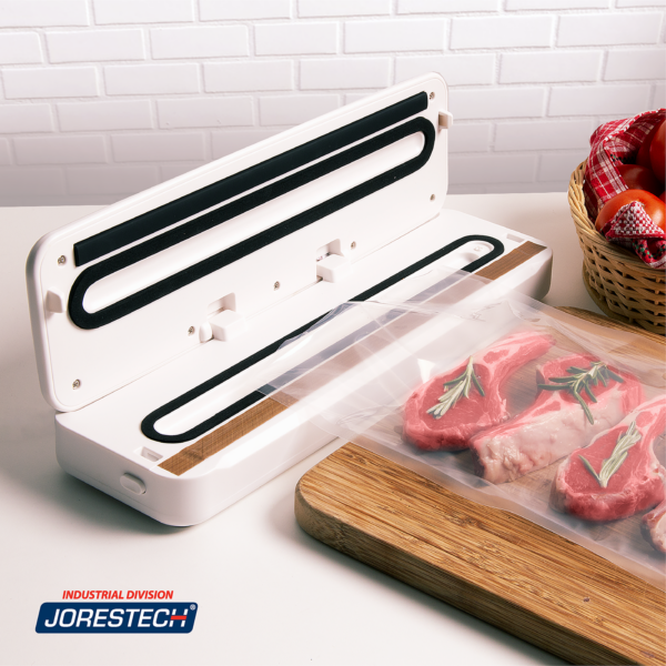 Máquina de sellado al vacío blanca JORES TECHNOLOGIES® sobre la encimera de una cocina doméstica. La máquina está junto a una bolsa al vacío abierta y llena de carne roja y romero, colocada sobre una tabla de cortar de madera, lista para ser sellada.