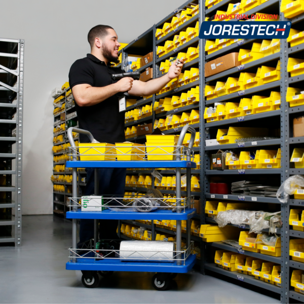 Un hombre en un almacén escaneando algunos artículos. Está usando el carrito utilitario de empuje JORES TECHNOLOGIES® de 3 estantes para transportar sus artículos por el lugar de trabajo.