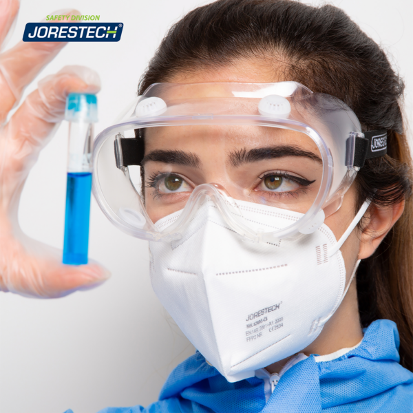 Mujer con gafas de seguridad ventiladas antivaho y ropa desechable en un entorno de laboratorio. Está inspeccionando un líquido azul dentro de un tubo de vidrio.